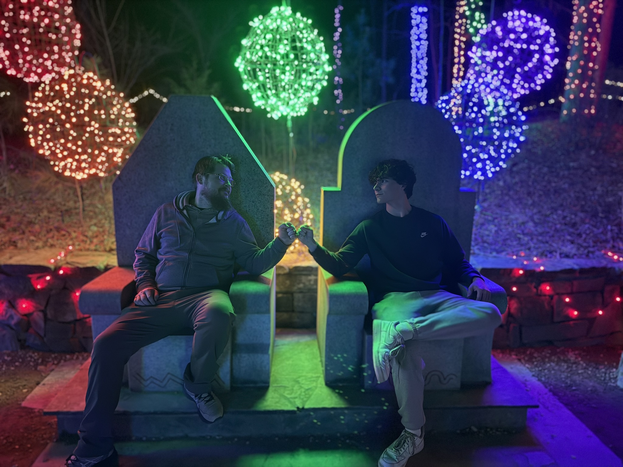 Ben and his son at a holiday light display, fist-bumping between two stone thrones lit in green, blue and red.